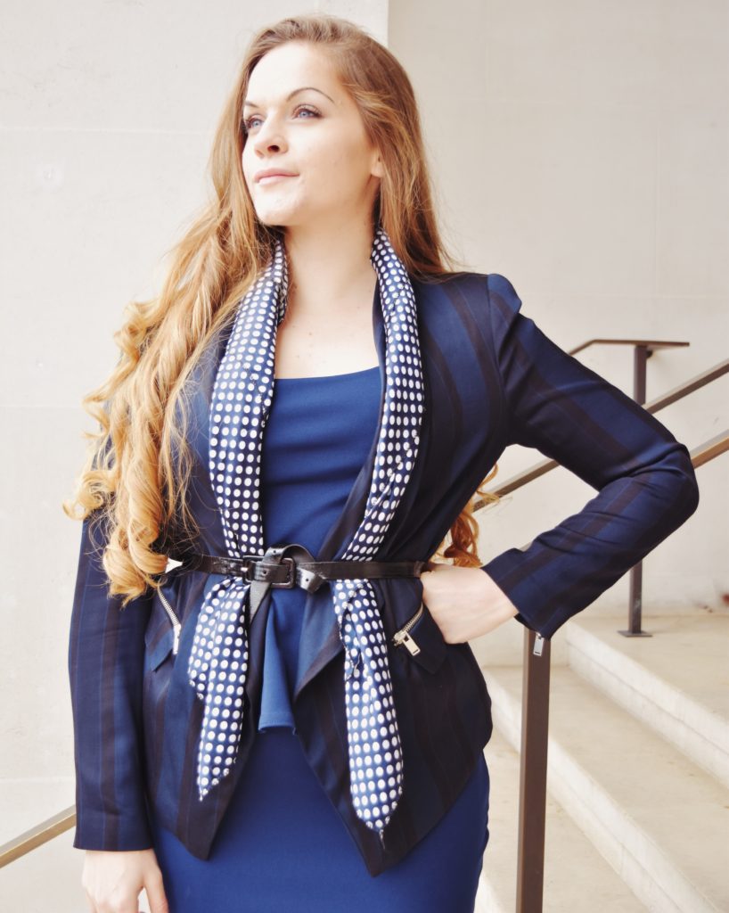 Power posing with head up and open arm wearing a smart blue suit and scarf